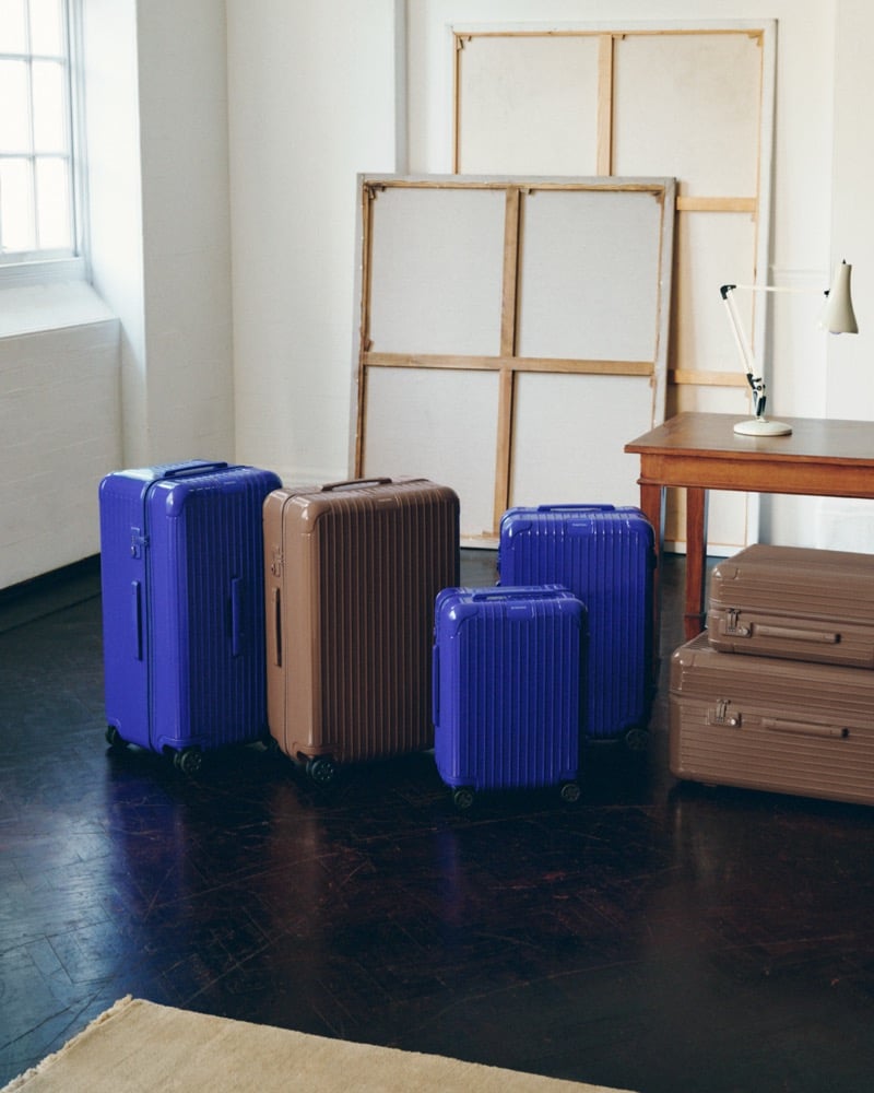 Five RIMOWA Essential suitcases in Ultramarine and brown standing in an artist's studio with canvases, a wooden table, and an architect lamp behind them