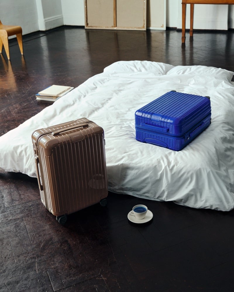 A brown RIMOWA cabin suitcase on the floor and an Ultramarine one on a white mattress, with a cup of blue liquid on a saucer and a stack of books nearby