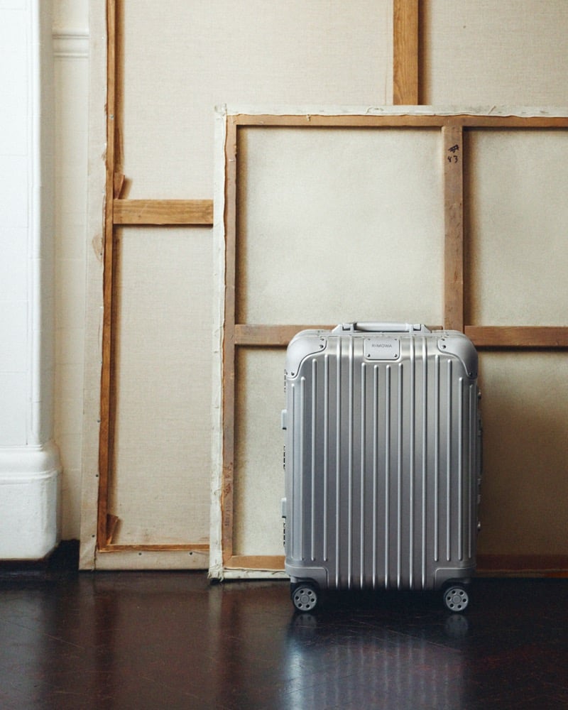 A silver aluminum RIMOWA cabin suitcase standing against a blank canvas frame on a dark wood floor