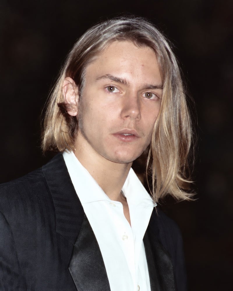 River Phoenix with shoulder-length, side-parted blonde hair, wearing a dark jacket over an open white shirt against a dark background.