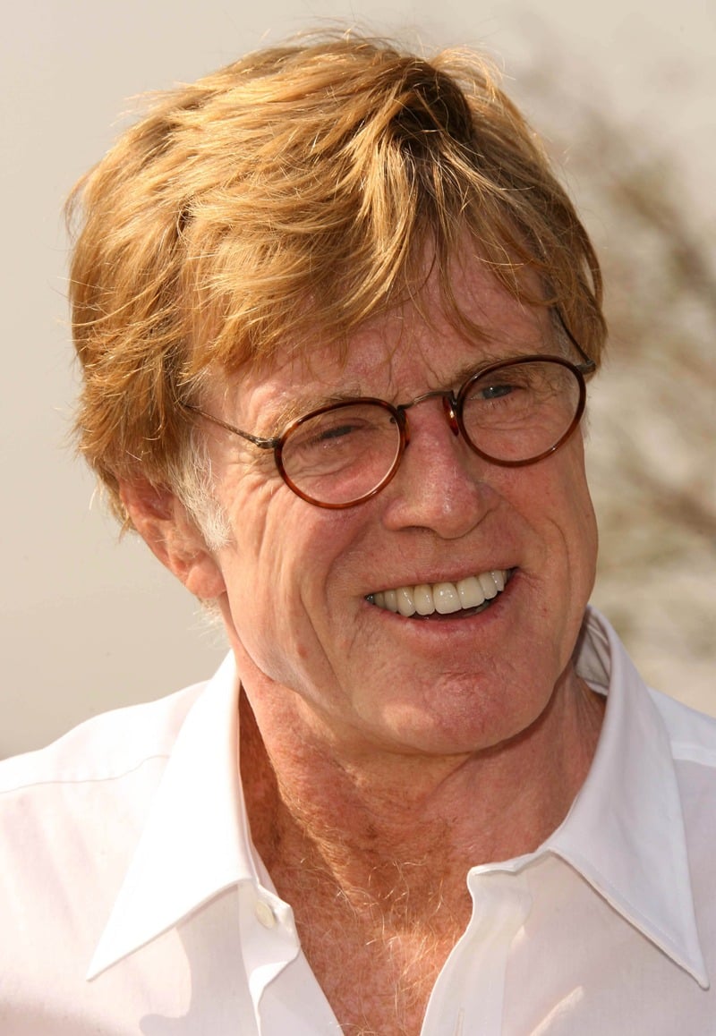 Robert Redford smiling with tousled golden hair and round dark-framed glasses, wearing an open-collar white shirt.