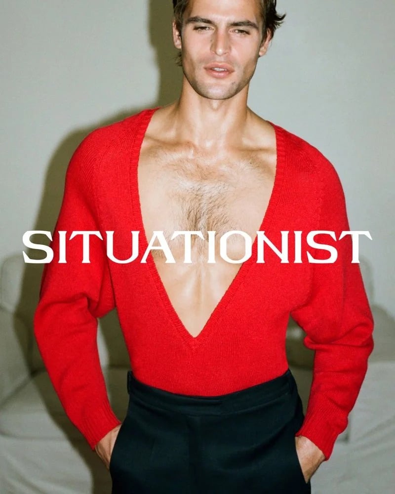 Parker van Noord wears a plunging red V-neck sweater and black trousers, with his hands in his pockets.