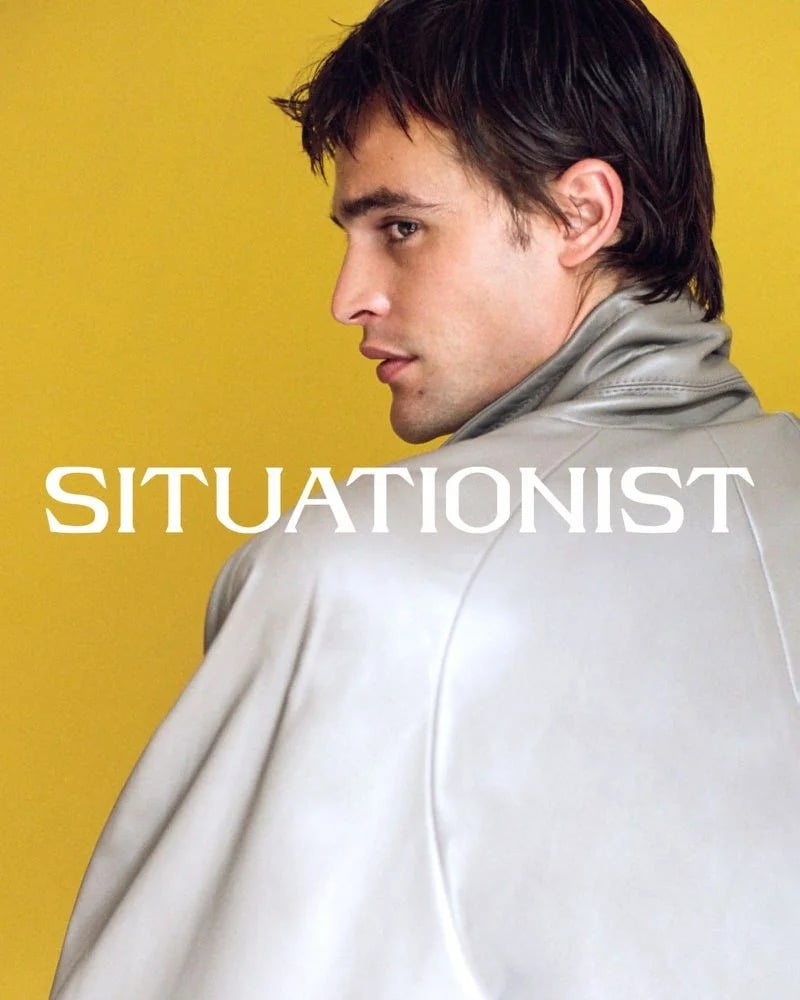 Parker van Noord looks over his shoulder in a gray high-collared jacket against a yellow wall.