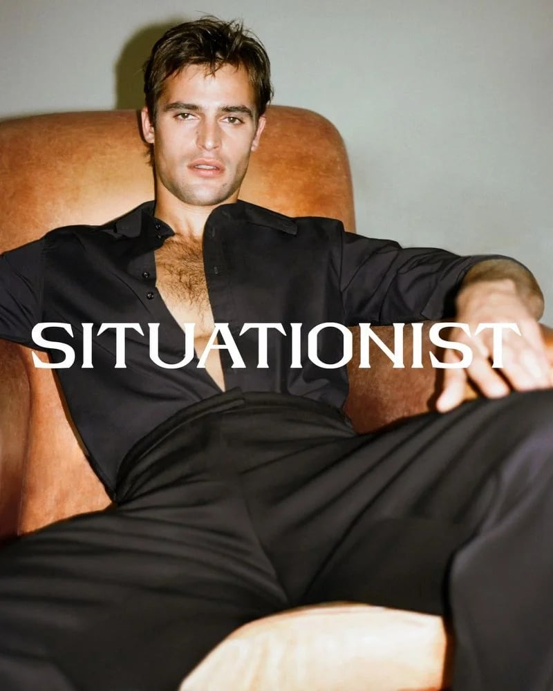 Parker van Noord reclines in a tan armchair wearing an open black shirt and black trousers.
