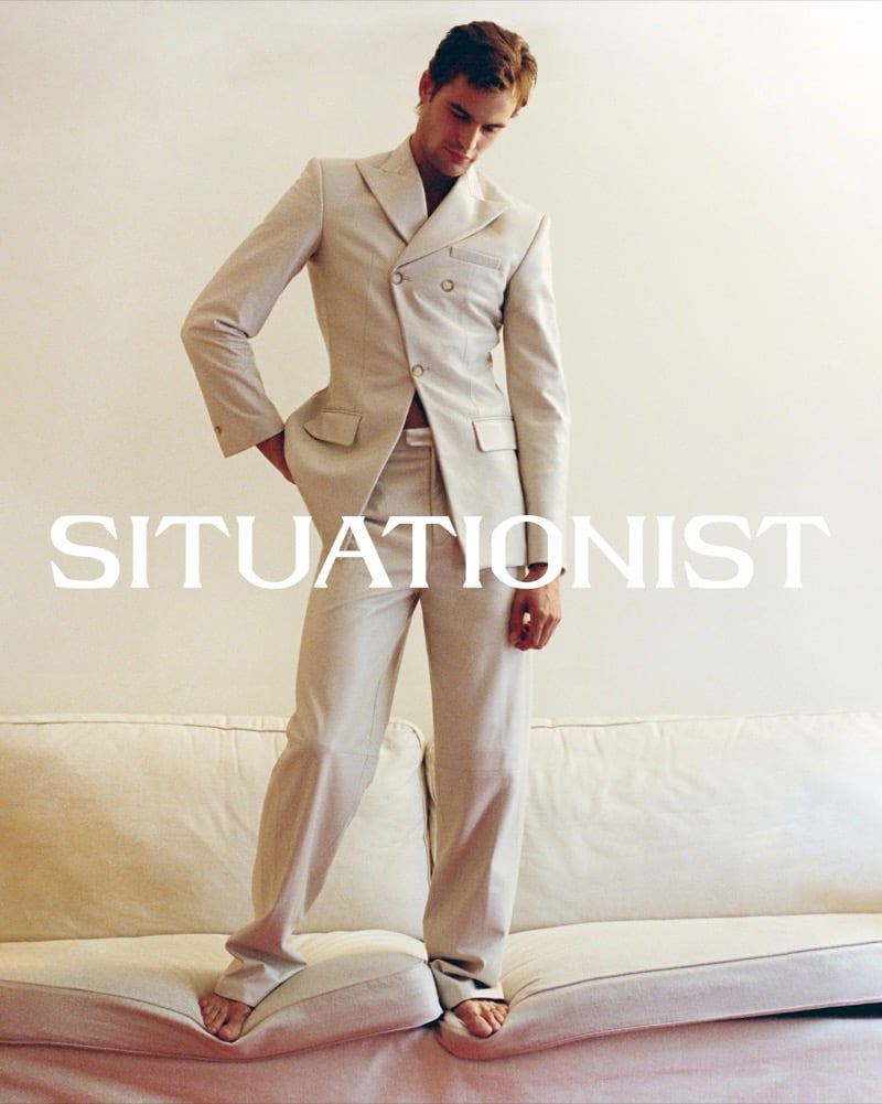 Parker van Noord stands barefoot on a cream sofa wearing a fitted cream double-breasted suit.