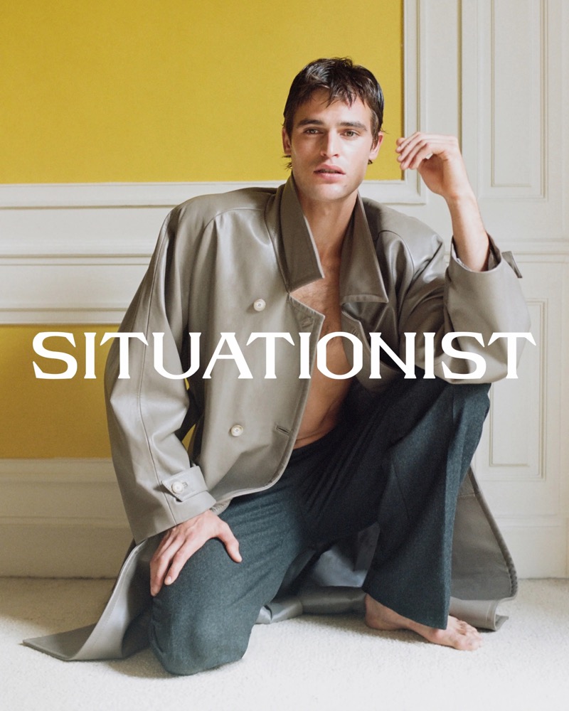 Parker van Noord kneels barefoot in an open gray coat and dark trousers against a yellow wall with white paneling.