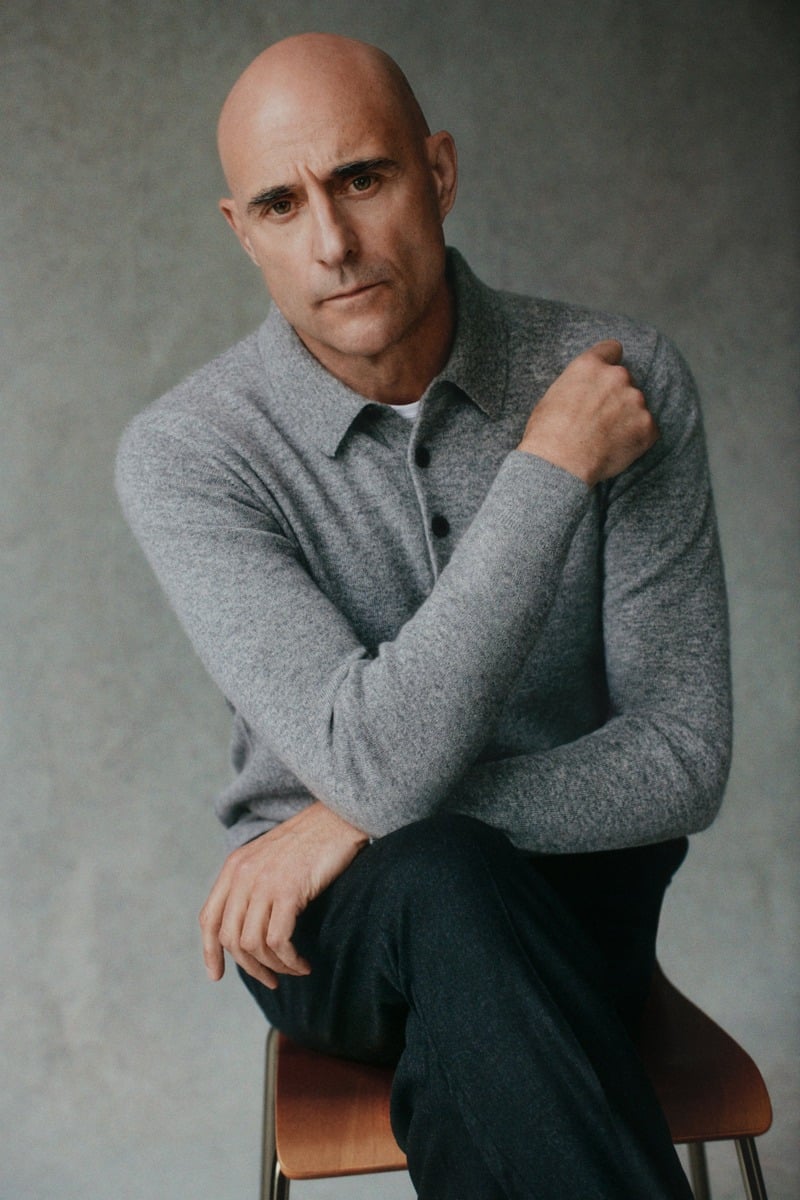 Mark Strong leans forward in a gray knit polo and dark trousers.