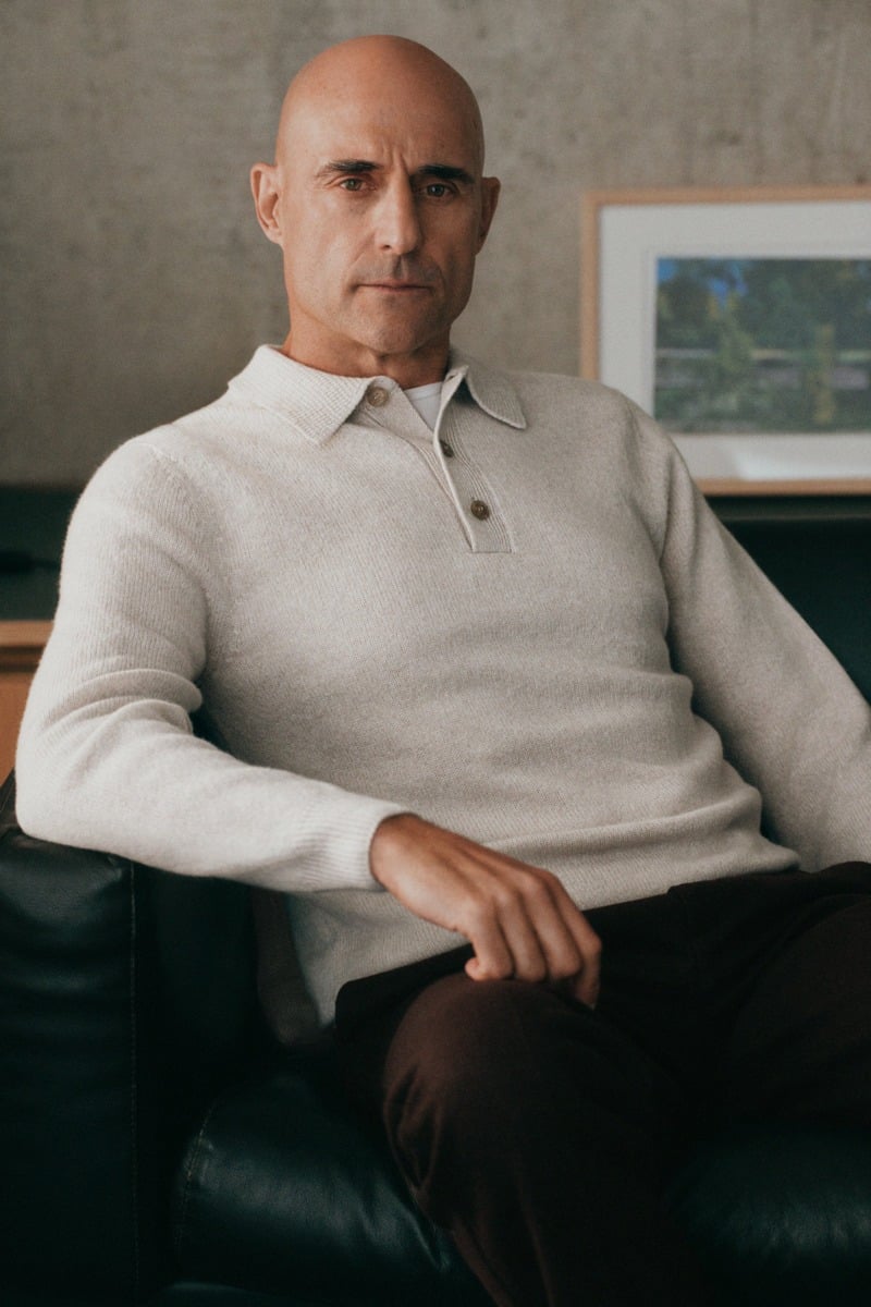 Mark Strong sits in an armchair wearing a cream long-sleeve knit polo and dark trousers.