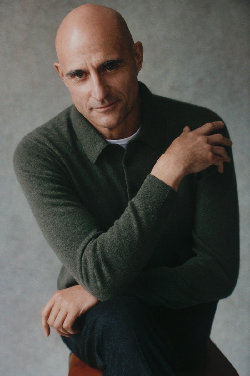 Mark Strong wears a dark green knit polo while seated on a stool.