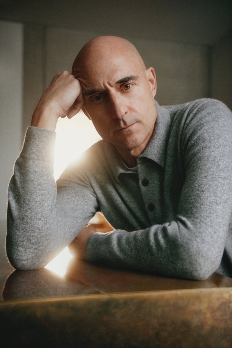 Mark Strong rests his head against his hand while wearing a gray knit polo.