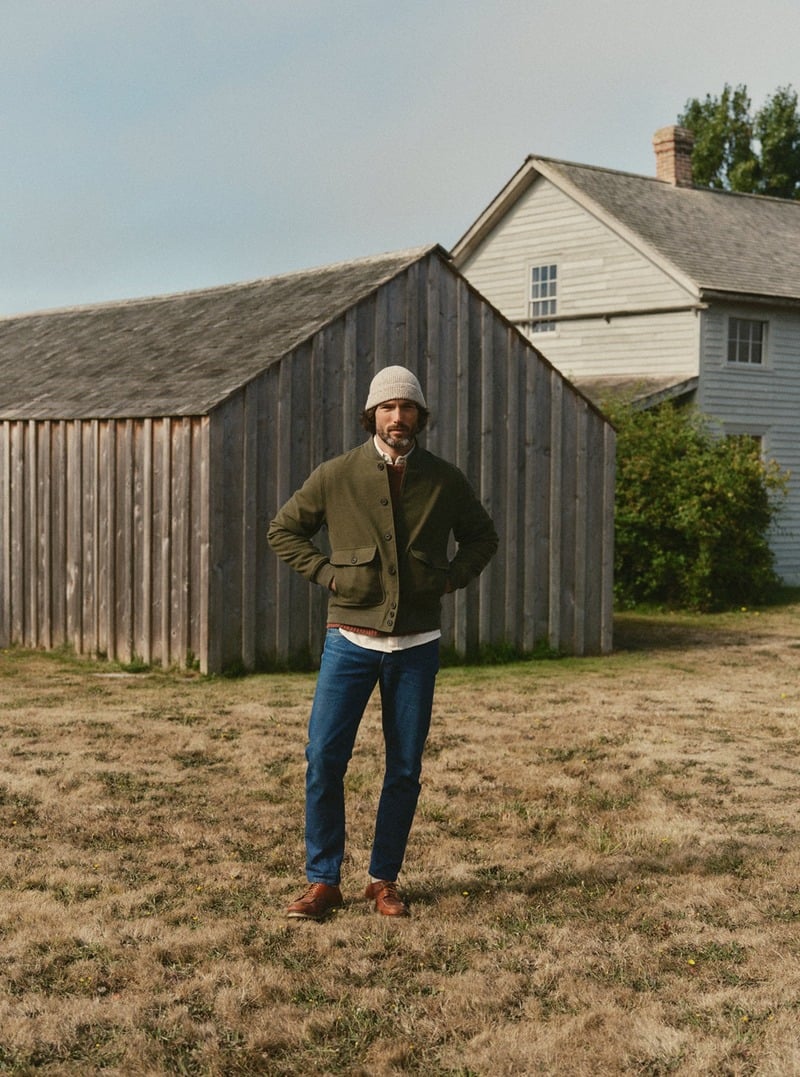 Model wearing an olive jacket with blue jeans beside a wooden barn. Taylor Stitch winter 2026 collection.