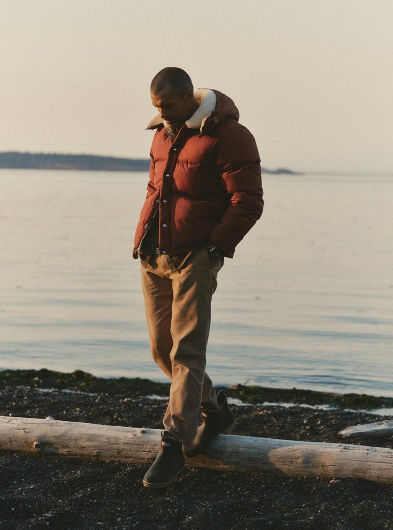 Model wearing a red down jacket and brown trousers on a rocky shoreline. Taylor Stitch winter 2026 collection.