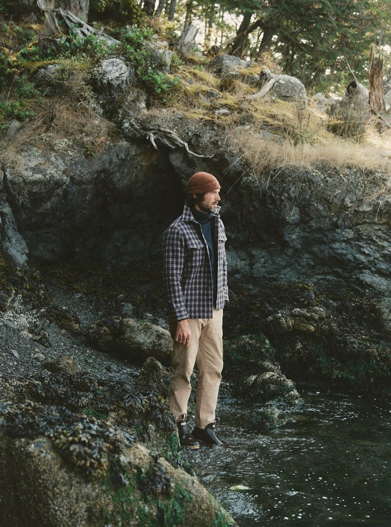 Model wearing a plaid shirt jacket and light trousers in a wooded landscape. Taylor Stitch winter 2026 collection.