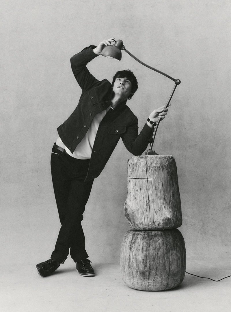 Black-and-white portrait of a model holding a lamp while wearing a trucker jacket and dark jeans. Taylor Stitch winter 2026 collection.
