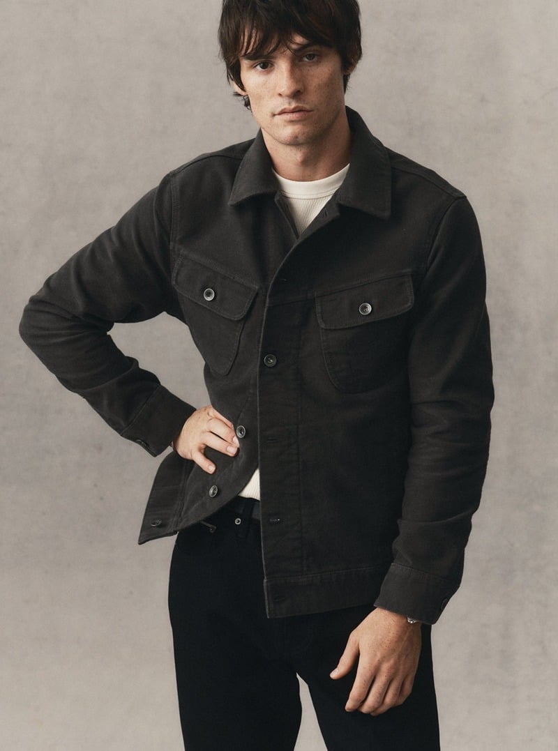 Model wearing a dark trucker jacket over a white T-shirt with dark jeans. Taylor Stitch winter 2026 collection.