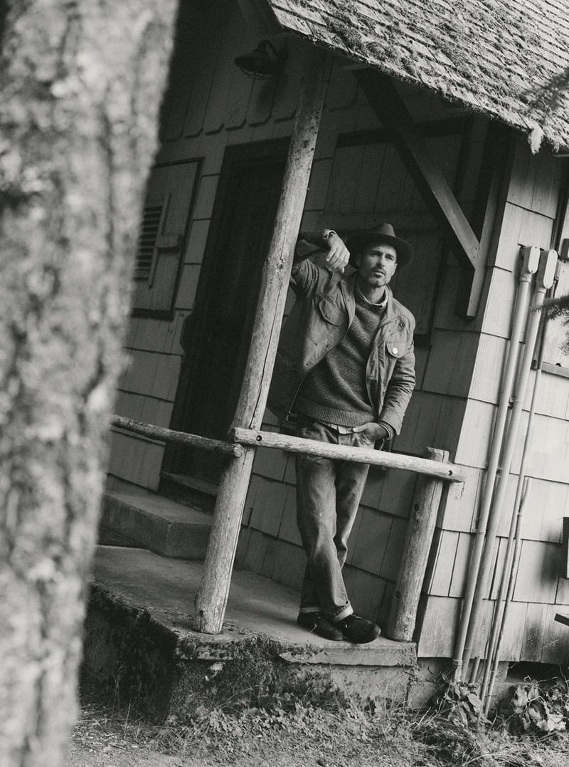 Black-and-white photograph of a model wearing a field jacket on a cabin porch. Taylor Stitch winter 2026 collection.