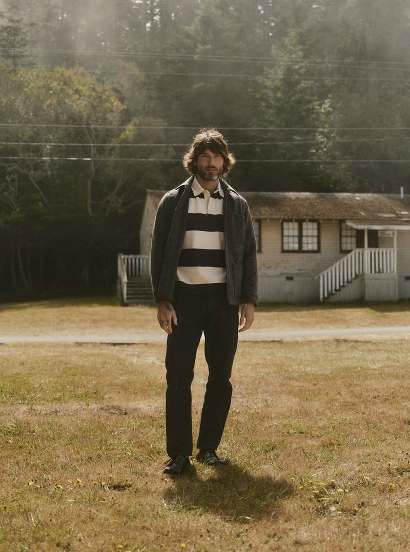 Model wearing a broad-striped rugby shirt with dark trousers in a field. Taylor Stitch winter 2026 collection.