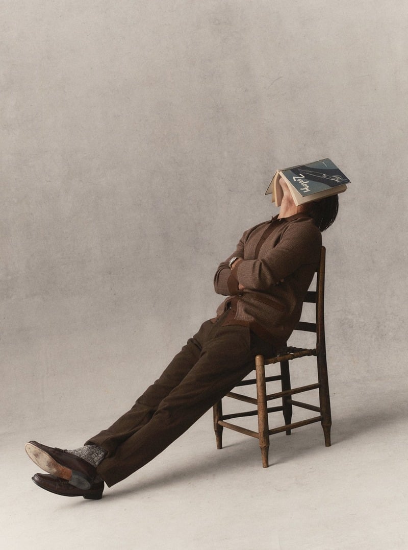 Model reclining in a chair in a brown sweater polo and trousers with a book over his face. Taylor Stitch winter 2026 collection.