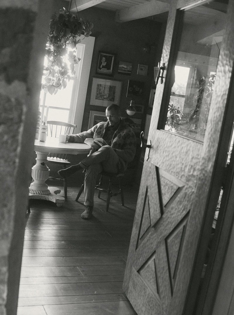 Black-and-white photograph of a man reading in a chair inside a wood-paneled room. Taylor Stitch winter 2026 collection.
