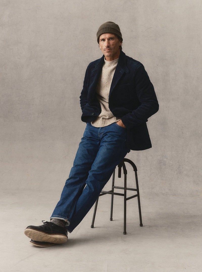 Model seated on a stool wearing a navy sportcoat over a pale sweater with blue jeans. Taylor Stitch winter 2026 collection.