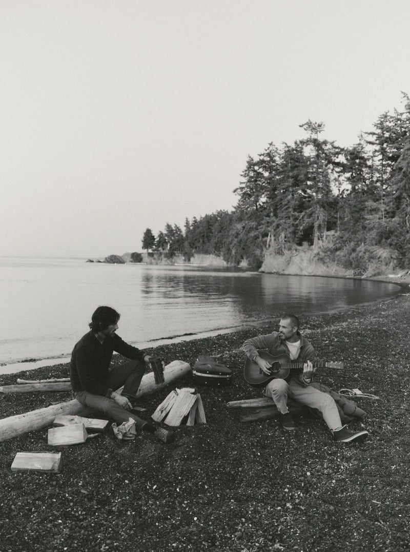 Black-and-white photograph of two men sitting by the shore with a guitar. Taylor Stitch winter 2026 collection.