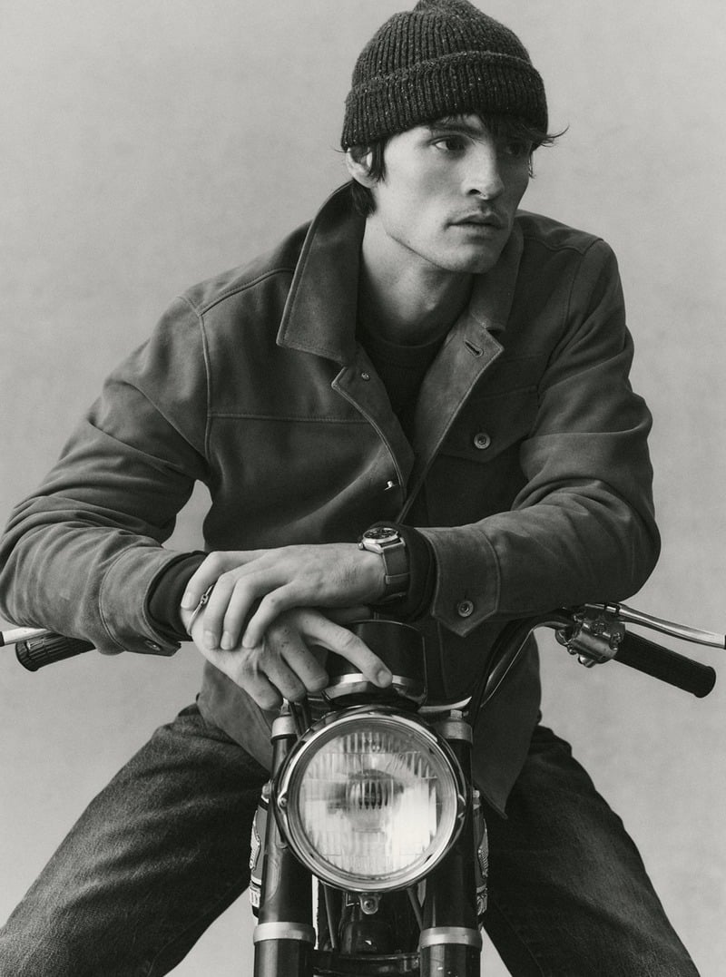 Black-and-white portrait of a model in a suede jacket and knit beanie sitting on a motorcycle. Taylor Stitch winter 2026 collection.