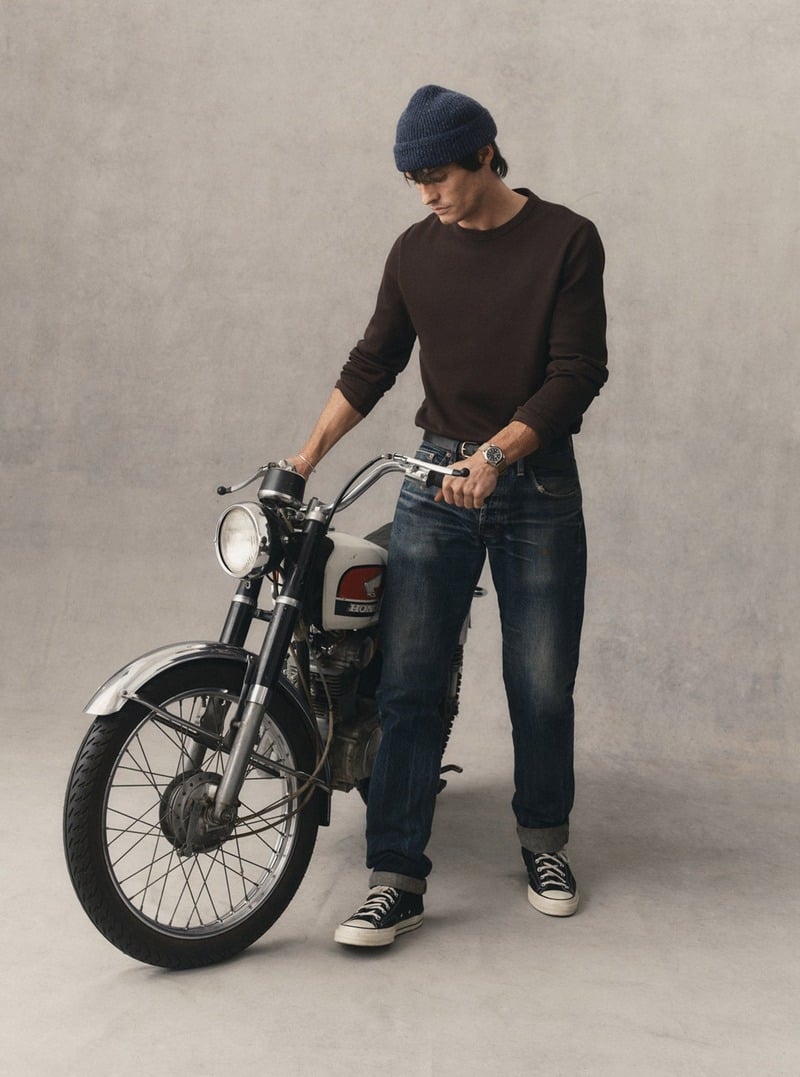 Model wearing a dark sweater with jeans and a knit beanie beside a vintage motorcycle. Taylor Stitch winter 2026 collection.