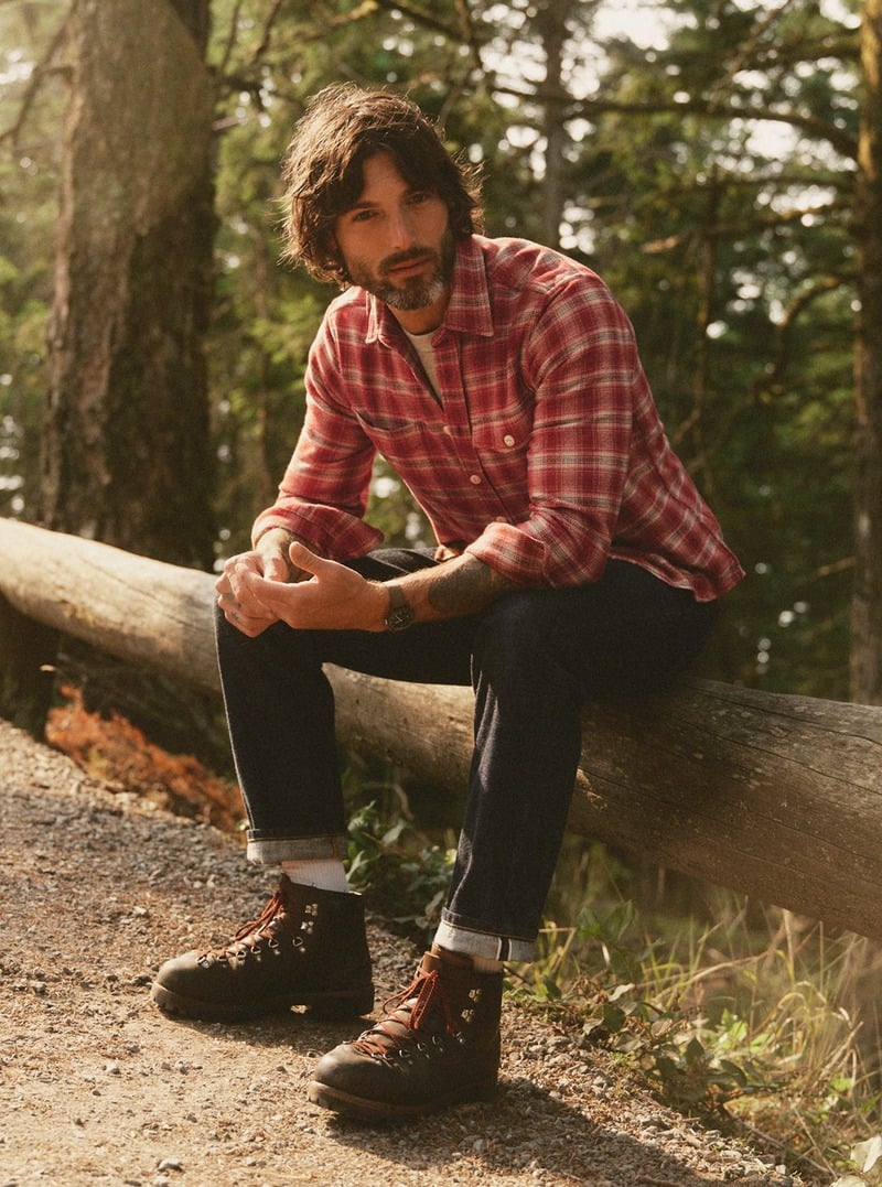 Model wearing a red plaid flannel shirt with cuffed dark jeans and boots while sitting on a log. Taylor Stitch winter 2026 collection.