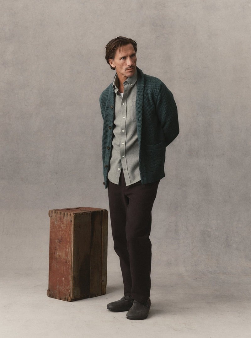 Model wearing a dark green shawl-collar cardigan over a gray button-down shirt with dark trousers. Taylor Stitch winter 2026 collection.