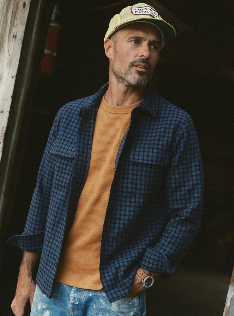 Model wearing a blue checked shirt over a mustard T-shirt with light blue jeans. Taylor Stitch winter 2026 collection.