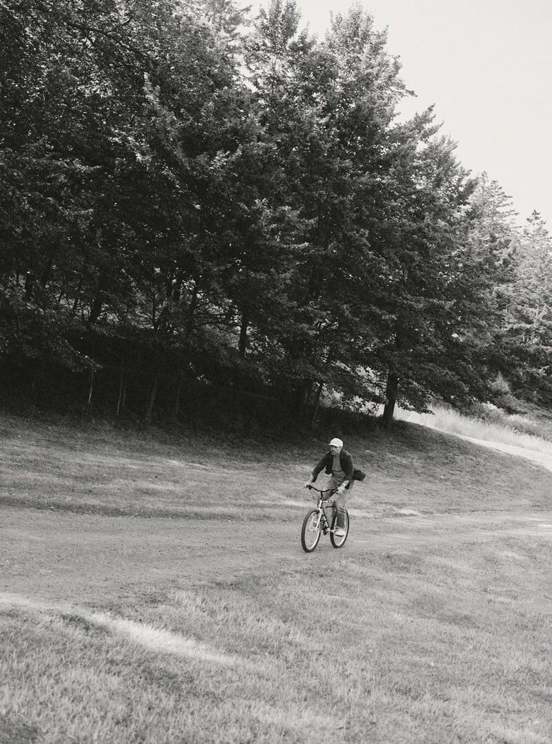 Black-and-white photograph of a man cycling along a wooded road. Taylor Stitch winter 2026 collection.
