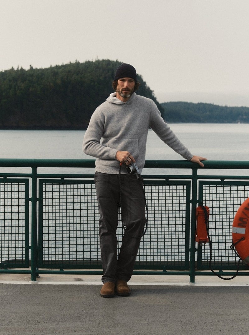 Model wearing a gray sweater hoodie with dark trousers beside the water. Taylor Stitch winter 2026 collection.