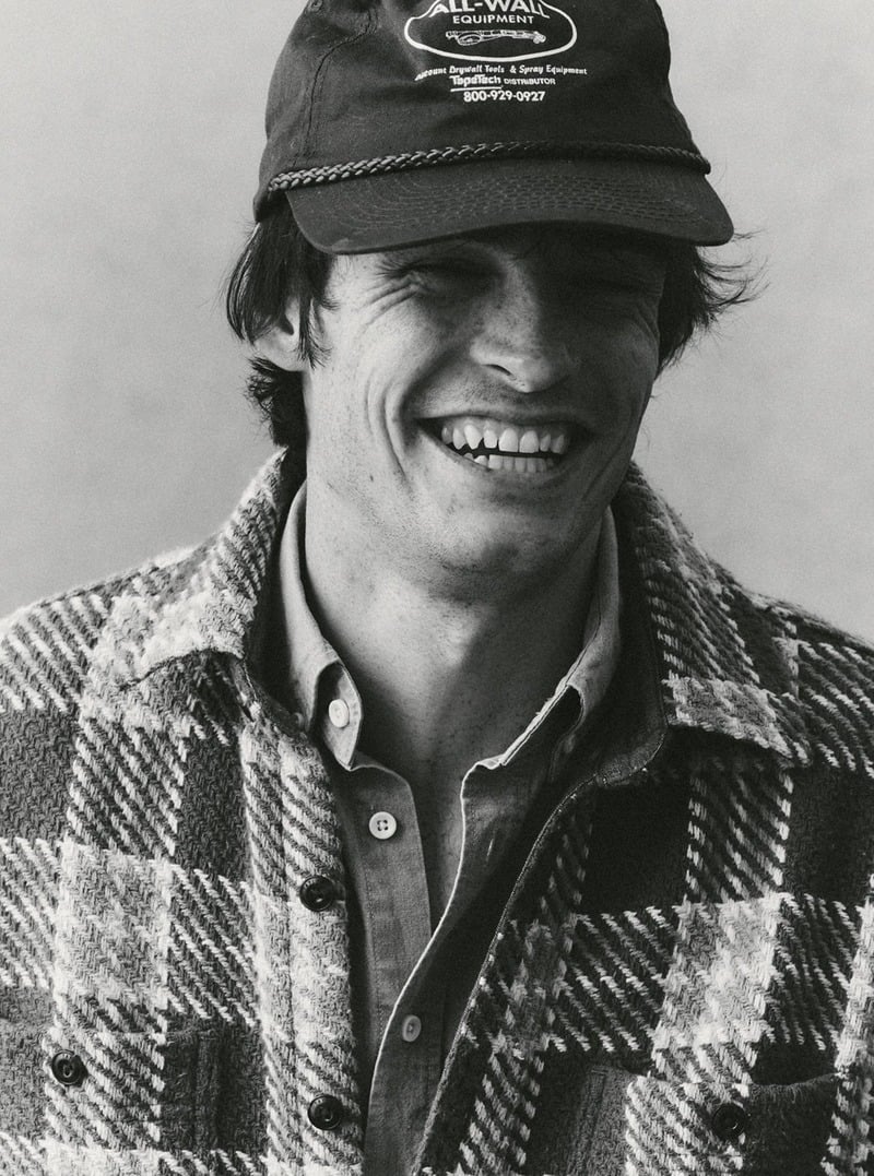 Black-and-white portrait of a smiling model wearing a plaid overshirt and a cap. Taylor Stitch winter 2026 collection.