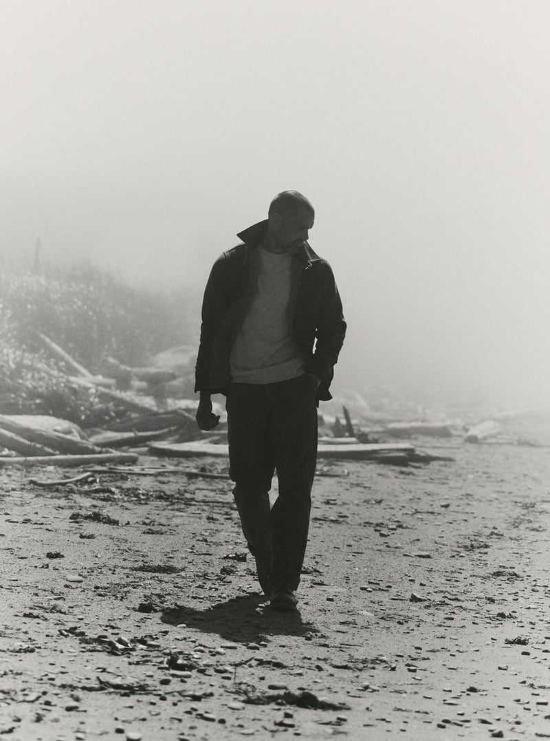 Black-and-white photograph of a model walking along a misty beach in a dark jacket. Taylor Stitch winter 2026 collection.