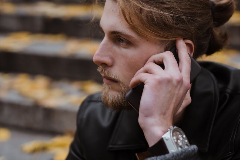 Close-up of a bearded man in a black leather jacket and silver watch holding a black phone to his ear.