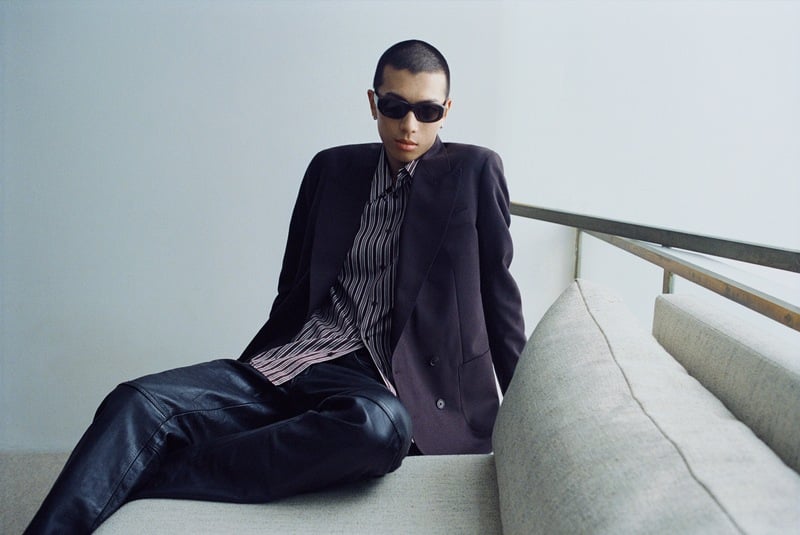 Yuyang Cai lounges on a sofa wearing dark sunglasses, a blazer, striped shirt, and dark trousers.