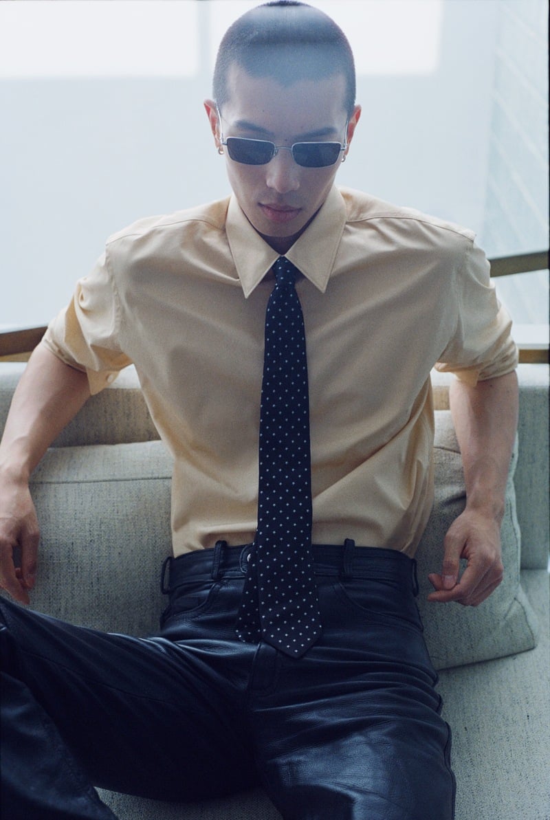 Yuyang Cai sits on a sofa in rectangular sunglasses, a beige shirt, dotted tie, and dark leather trousers.