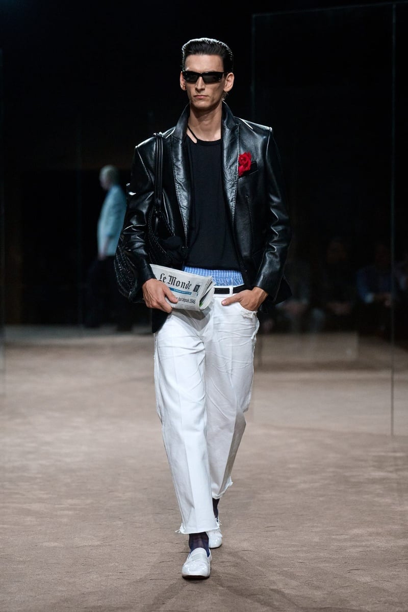 A model wears glossy dark jacket with a red rose, blue shirt, and white trousers on the Tom Ford spring-summer 2027 runway.