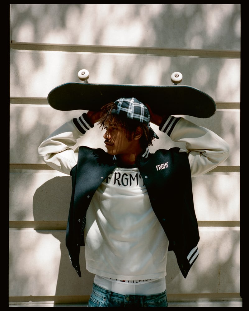 Nile Winchester holds a skateboard above his head in a varsity jacket, graphic T-shirt, and plaid cap.