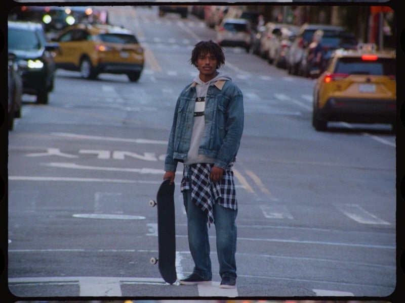 Nile Winchester stands with a skateboard in a denim jacket, relaxed jeans, and a plaid shirt tied at his waist.