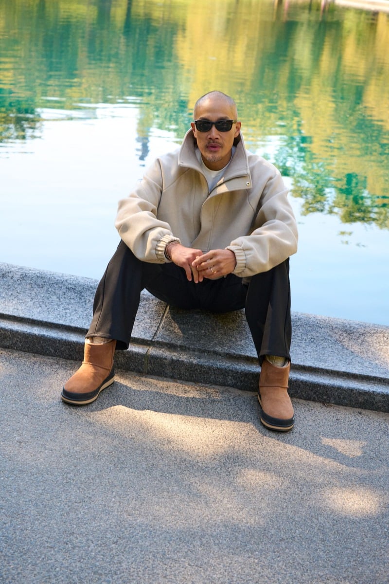 Dao-Yi Chow sits beside the water in a light quarter-zip, black trousers, and chestnut UGG Classic Mini Guard GTX boots.