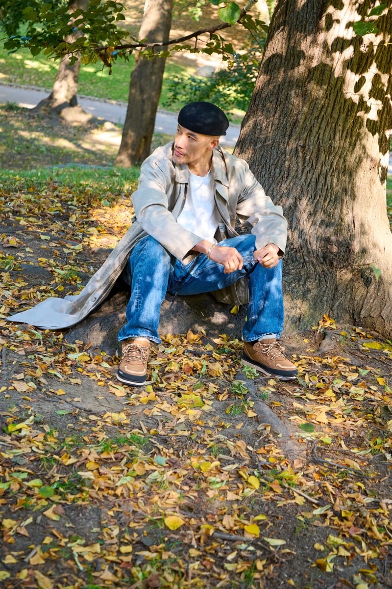 Dao-Yi Chow sits beneath a tree in a long trench coat, blue jeans, and chestnut UGG Lowmel Guard GTX shoes.