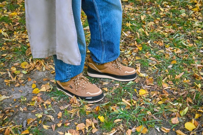 Dao-Yi Chow wears blue jeans over chestnut UGG Lowmel Guard GTX shoes on a leaf-covered path.
