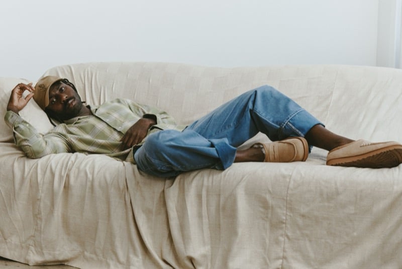 Dev Hynes reclines on a sofa in a plaid shirt, rolled-up jeans, and Chestnut UGG x JW Anderson loafers.