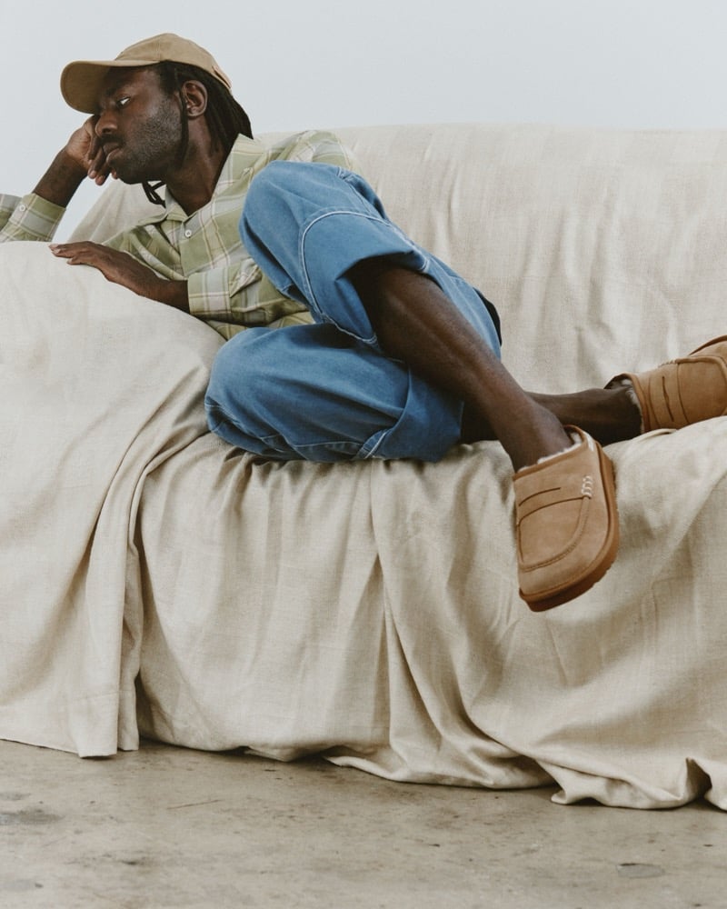 Dev Hynes lounges on a sofa in a hat, a striped shirt, jeans, and Chestnut UGG x JW Anderson loafers.