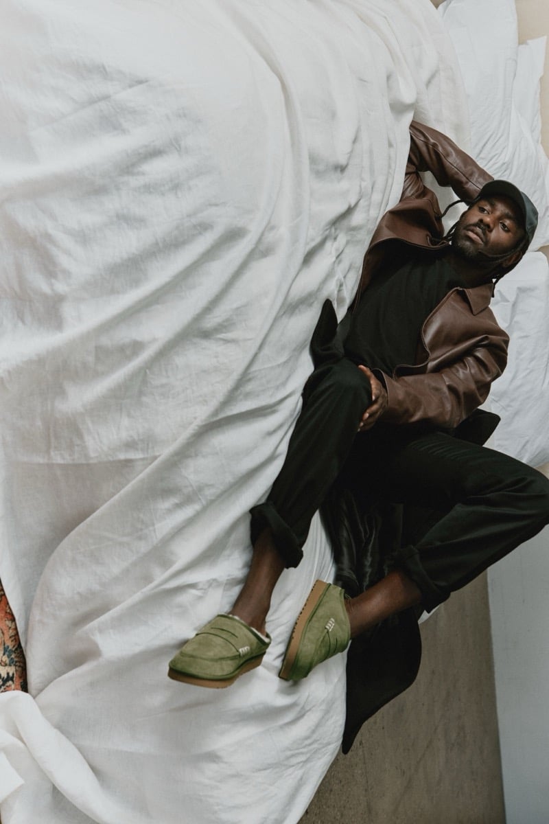 Dev Hynes reclines beside a draped sofa in a brown jacket, dark trousers, and Khaki UGG x JW Anderson loafers.