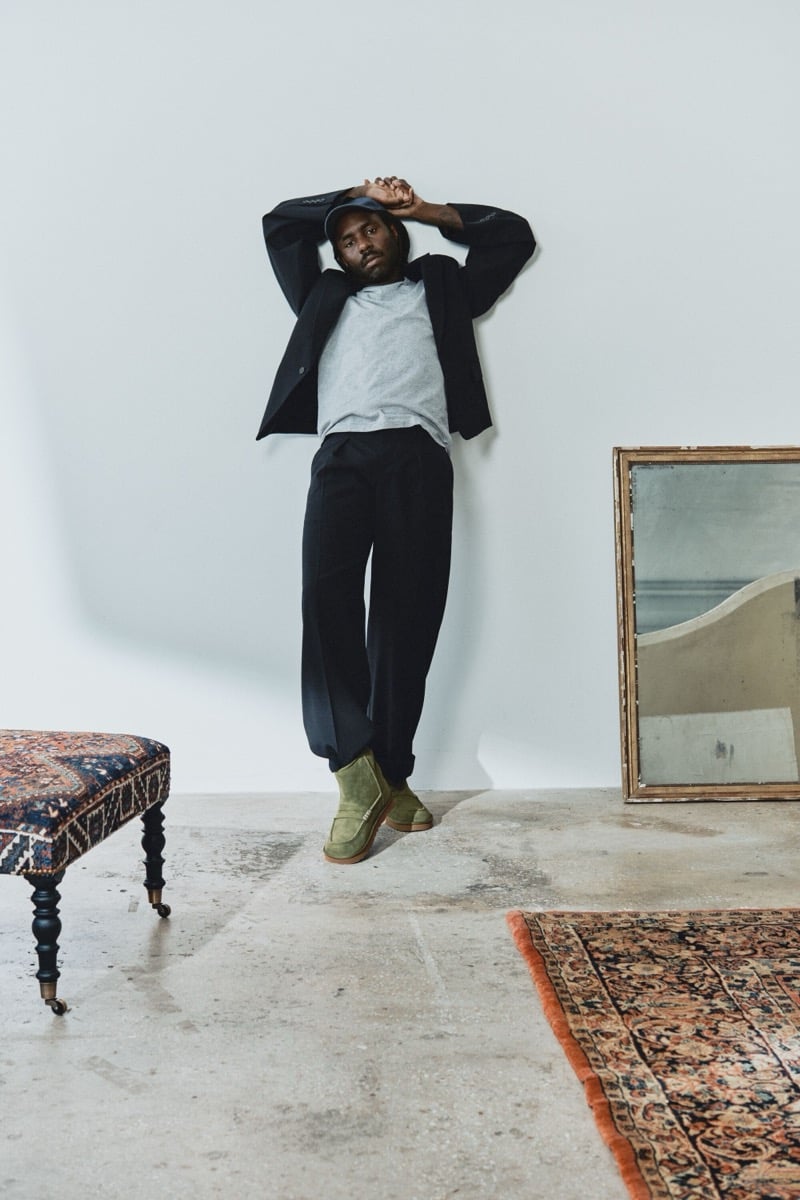 Dev Hynes poses in black tailoring and Khaki UGG x JW Anderson loafers with his arms raised.