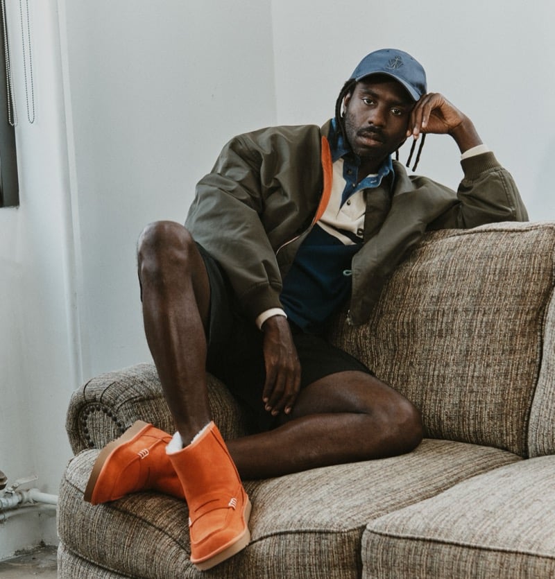Dev Hynes sits in an armchair wearing a blue cap, a jacket, shorts, and Orange UGG x JW Anderson loafer boots.