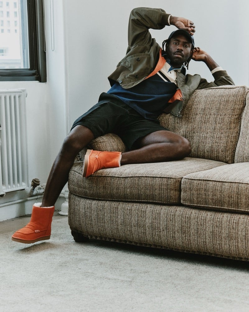 Dev Hynes reclines in an armchair wearing a jacket, shorts, and Orange UGG x JW Anderson loafer boots.