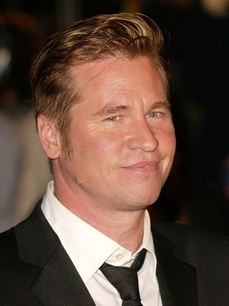 Val Kilmer with swept-back dark blonde hair and short sides, wearing a black suit, white shirt, and loosely knotted black tie.